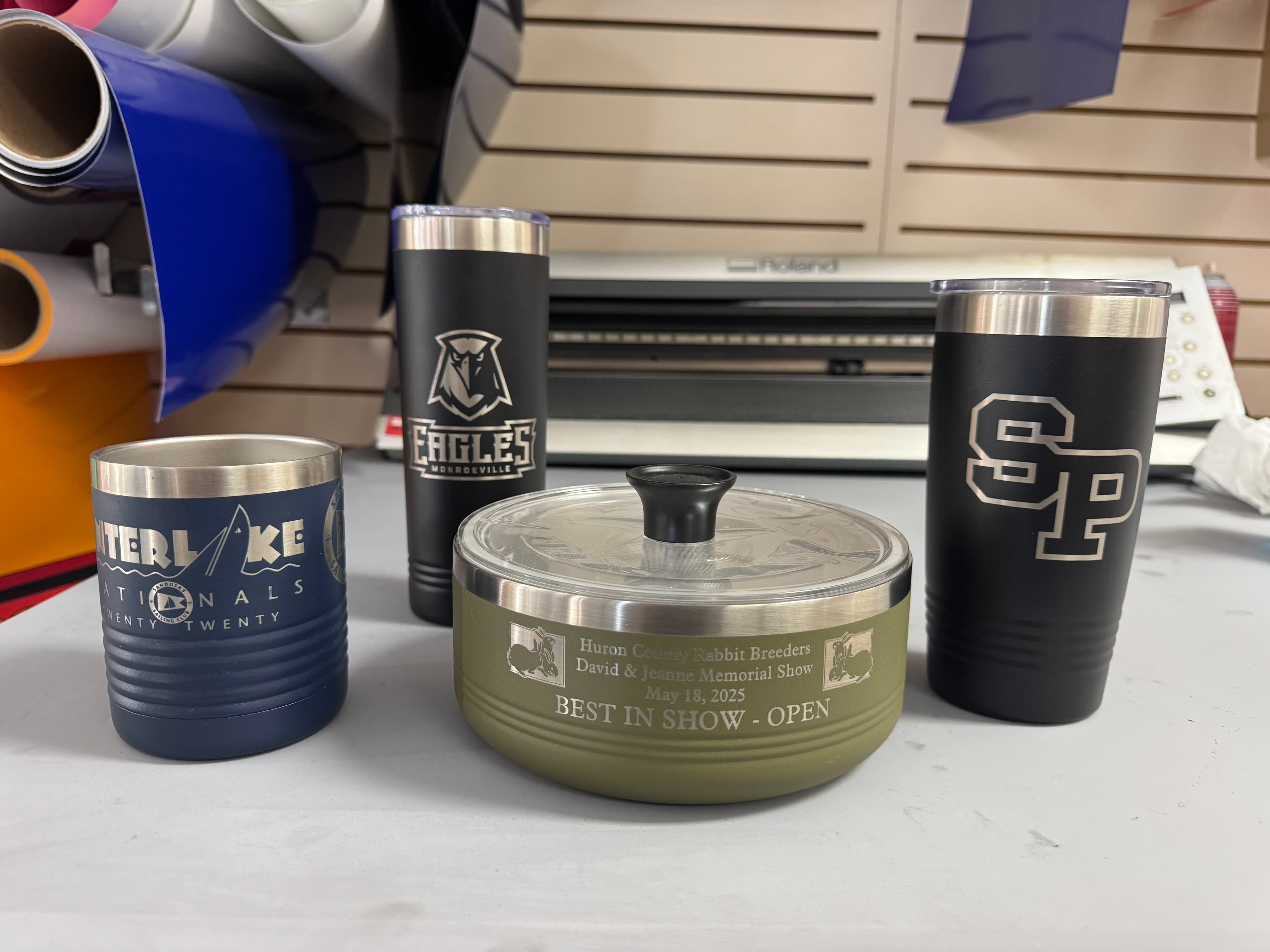 Three custom engraved insulated tumblers and a green lidded bowl on a workshop table.