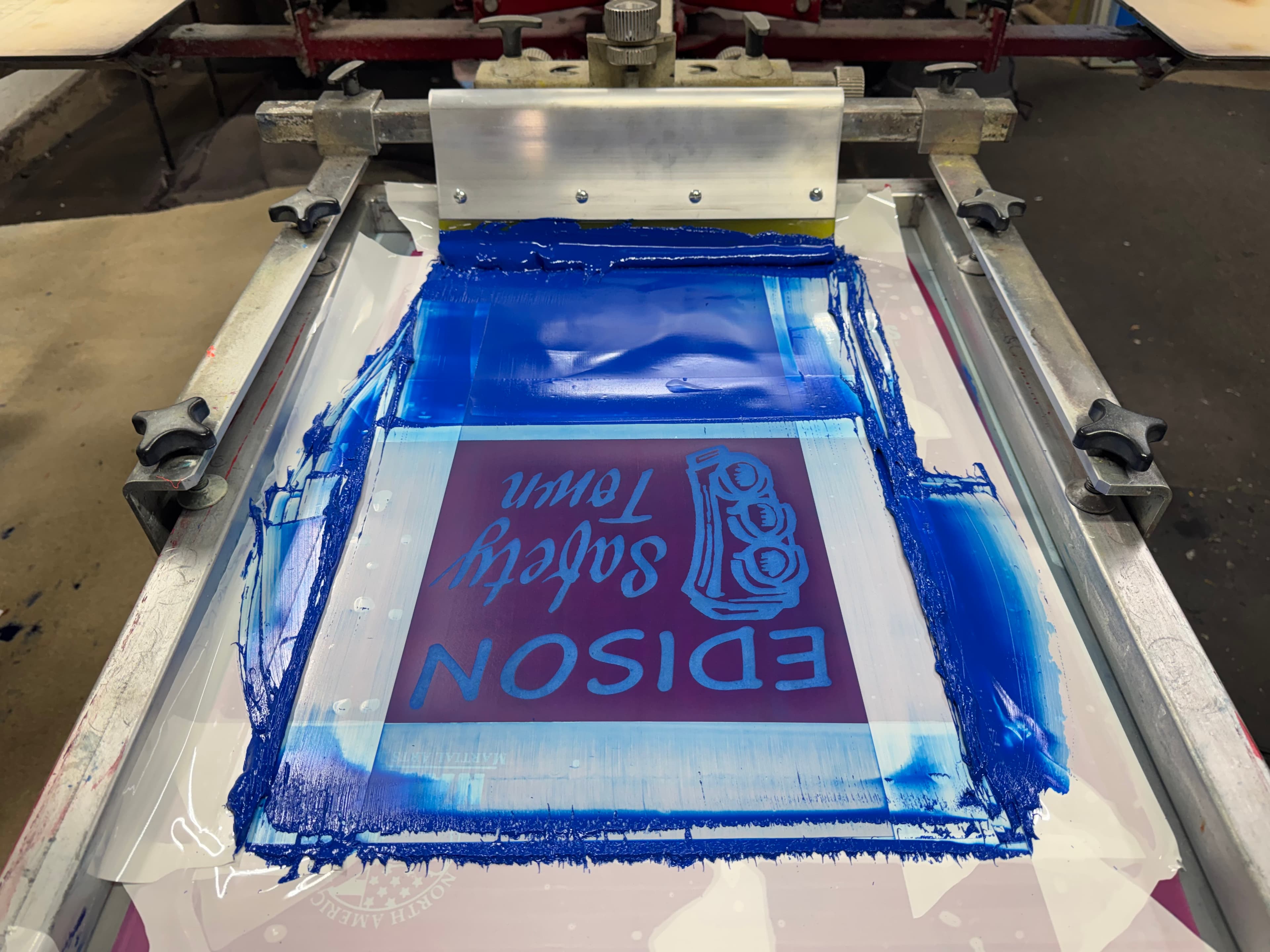 Screen printing press with blue ink over a reversed stencil reading EDISON Safety Town.