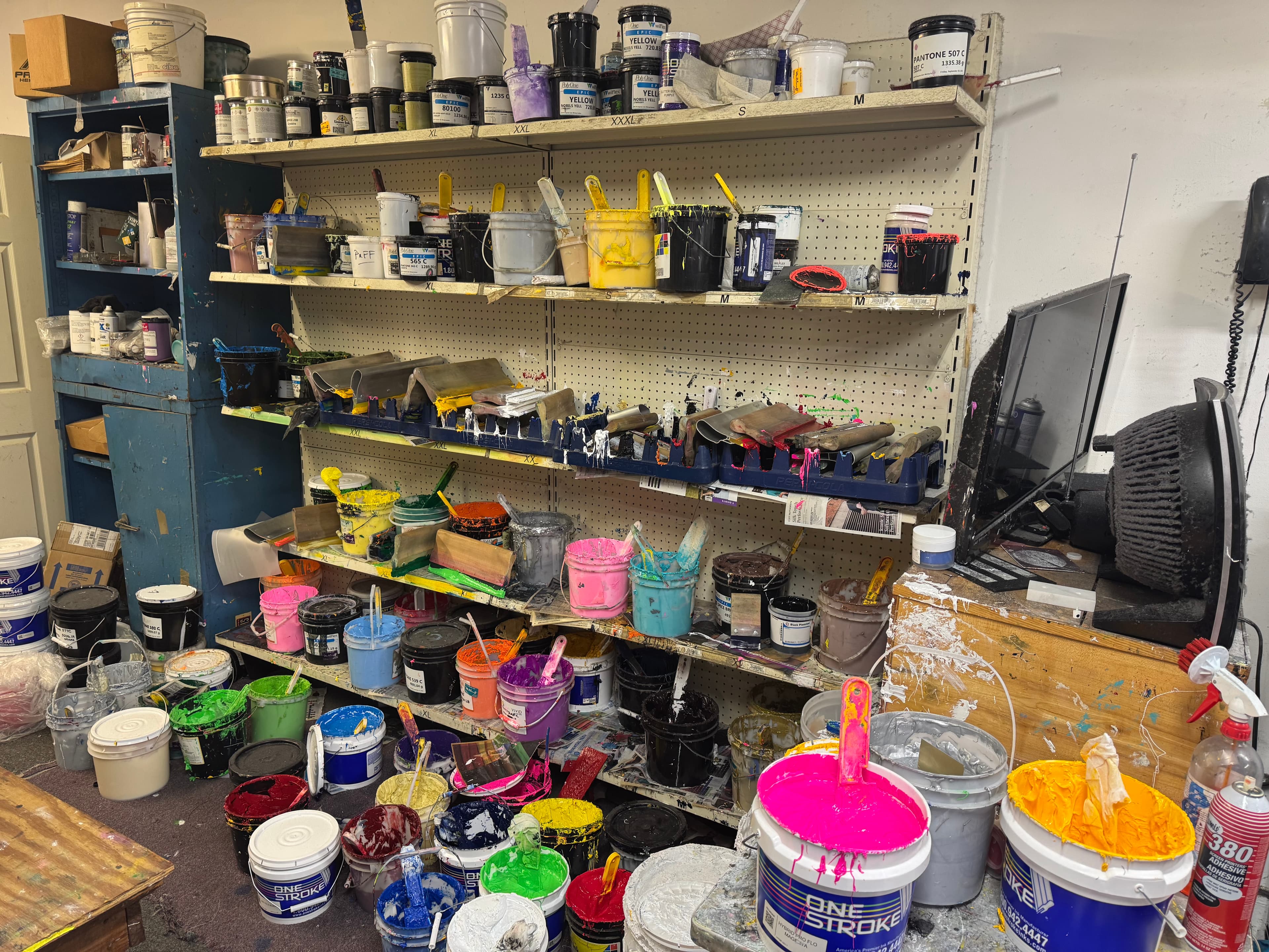 Shelves in a cluttered workshop packed with vibrant screen printing ink buckets and squeegee tools.