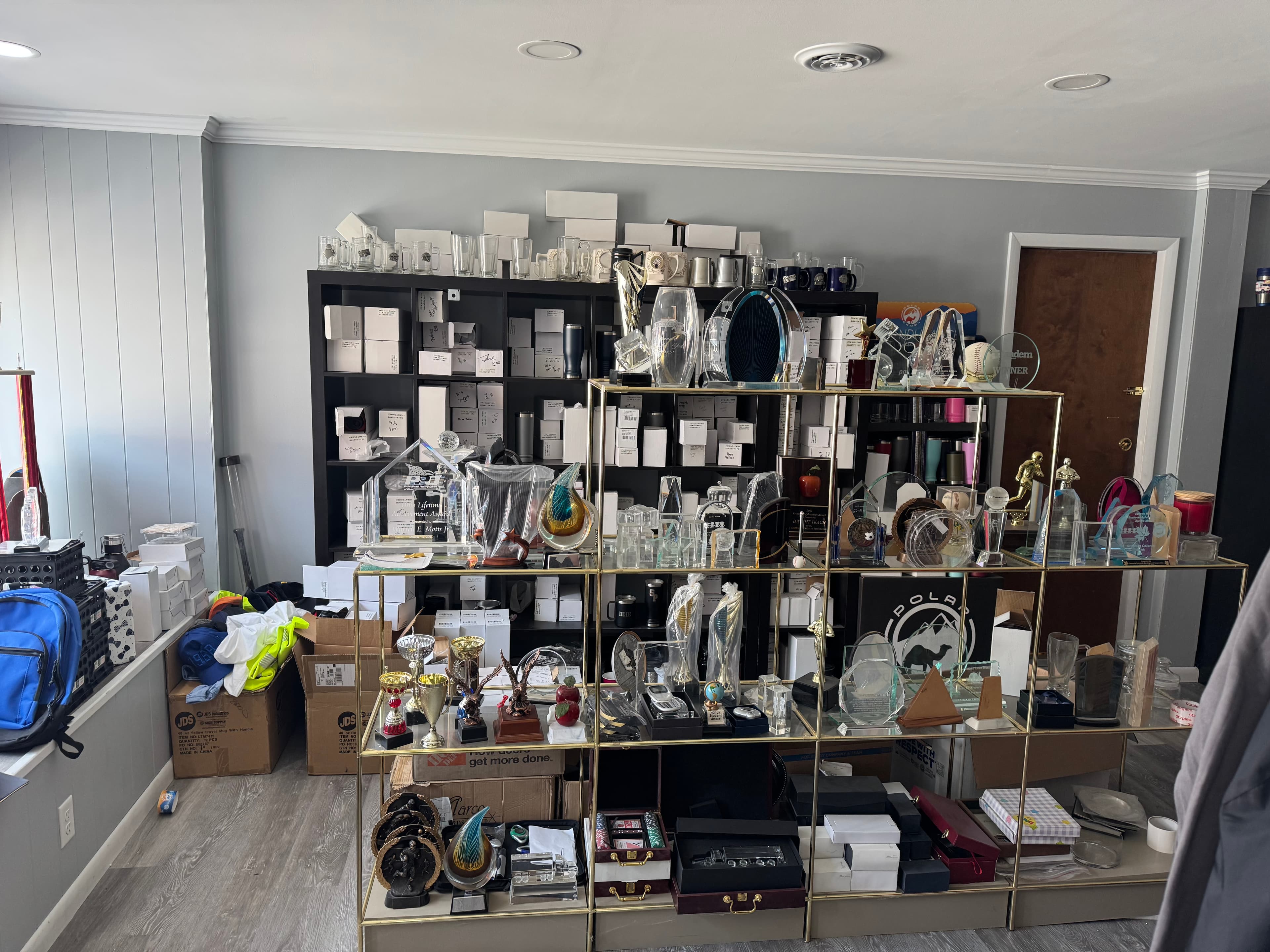 Gold and black shelves overflowing with trophies, glass awards, and boxes in a cluttered room.