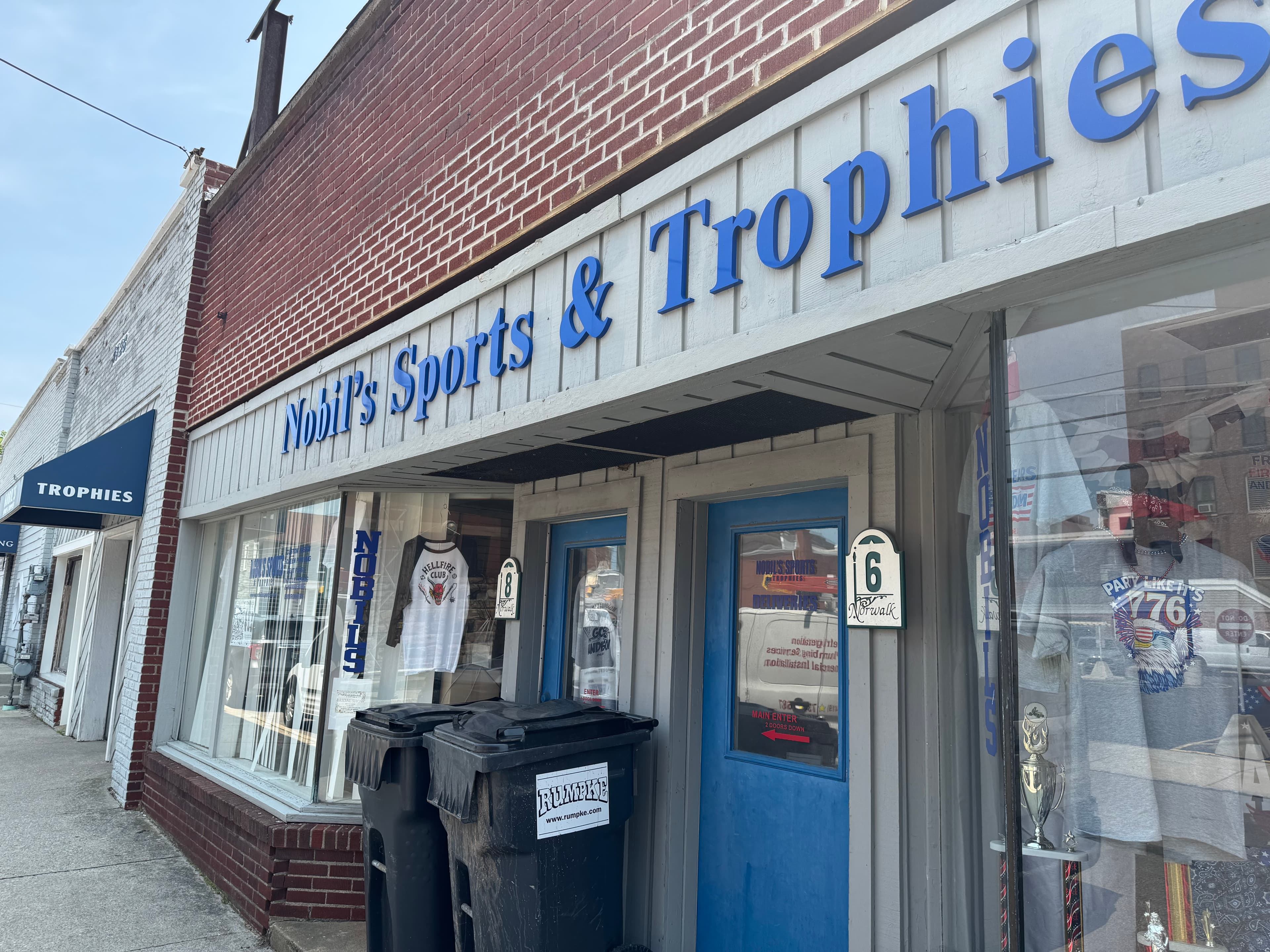 Storefront of Nobil's Sports & Trophies with blue signage, t-shirt displays, and blue entrance doors.