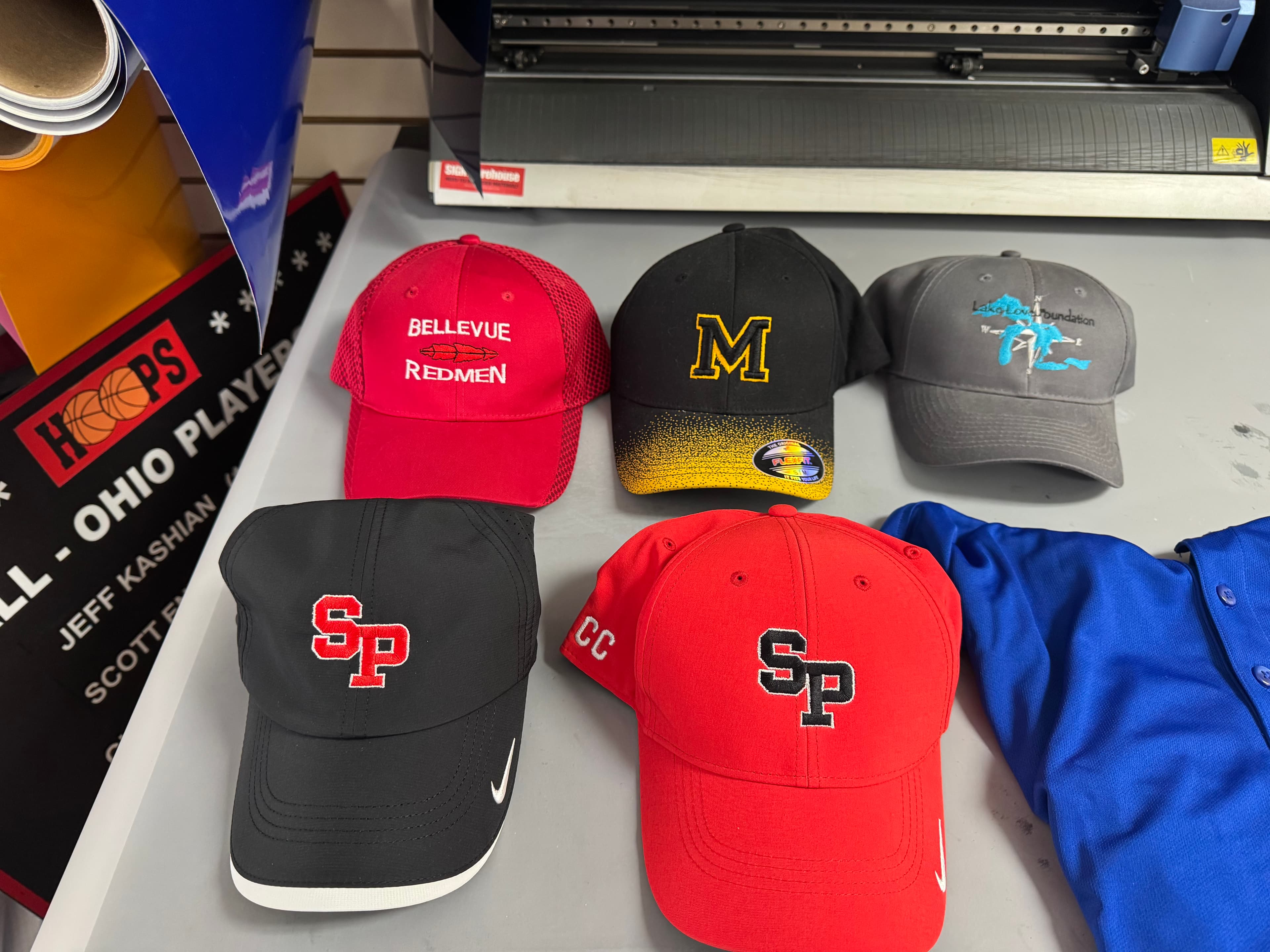 Five custom embroidered baseball caps with various logos displayed on a gray work table.