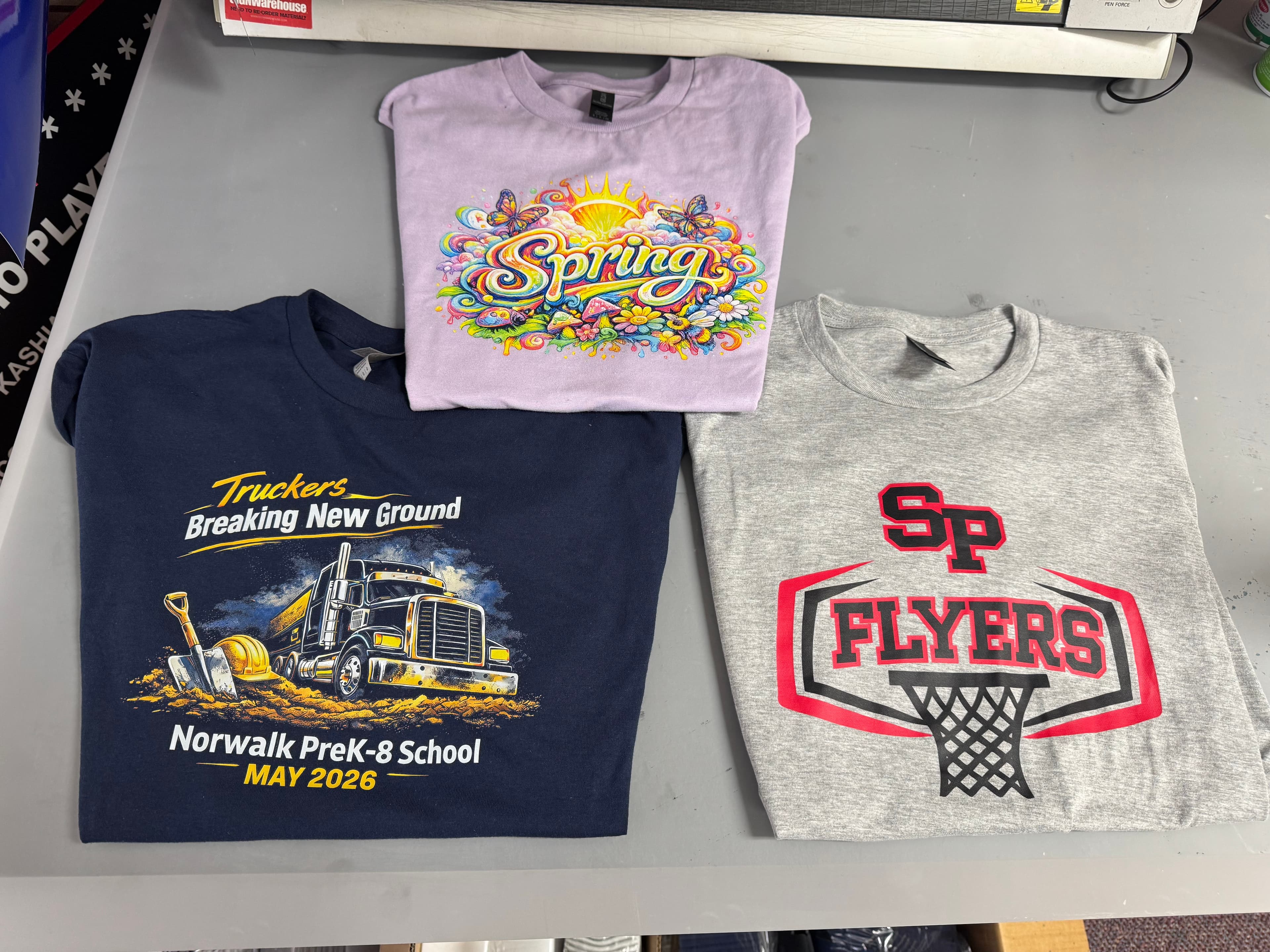 Three t-shirts in purple, navy, and grey featuring spring, school, and basketball team graphics.